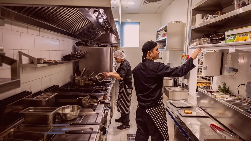 Cozinha profissional mostrando chefs em ação, um preparando alimentos e outro organizando utensílios, destacando a eficiência do trabalho em equipe em um ambiente de cozinha.