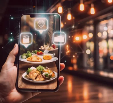 Smartphone exibindo pratos de comida em um ambiente de restaurante iluminado. O uso da tecnologia para compartilhar experiências gastronômicas é evidenciado por elementos virtuais.