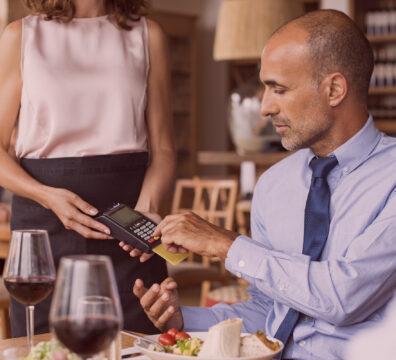 Homem pagando com cartão de crédito em restaurante elegante, enquanto a garçonete segura o terminal de pagamento, ao lado de taças de vinho e pratos de comida.